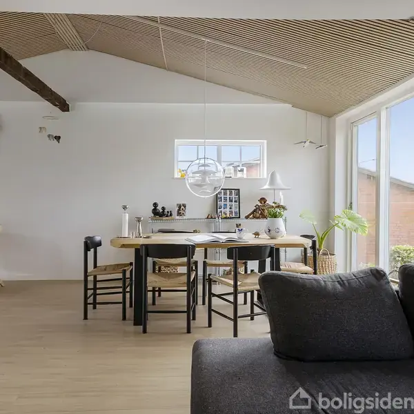 Sofa og spisebord i en stue med hvide vægge og lofter med træbjælker. Store vinduer bringer lys ind, og der er planter og kunst på væggene.