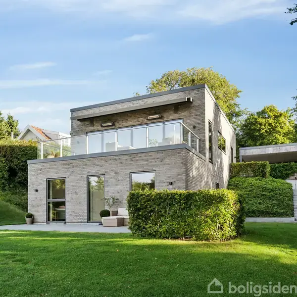 En moderne, to-etagers murstensvilla med store vinduer står foran en velholdt græsplæne. Omgivet af grønne buske og træer under en klar blå himmel.