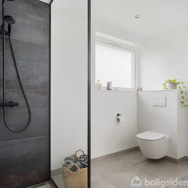 Brusekabine med sort bruser og væg, kurv med håndklæder; væghængt toilet under et vindue, en plante på væggen; lyst og minimalistisk badeværelse.