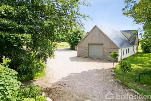 En stor garagebygning står stille omgivet af en grusvej og grøn vegetation. Træer skaber en naturlig baggrund i det solrige landskab.