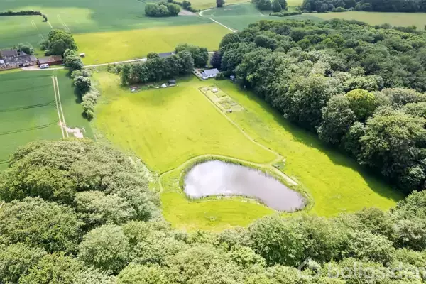 En lille sø ligger i en lysning omgivet af tæt skov og åbne græsmarker. En gård ses i baggrunden ved siden af små bygninger og en grusvej.