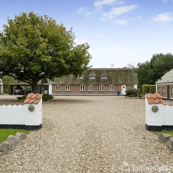 En gård med stråtag står stille, omkranset af grusvej og grønne træer, mellem to hvidmalede portstolper. Bygningerne er i traditionel, dansk landstil med røde murstensfacader.