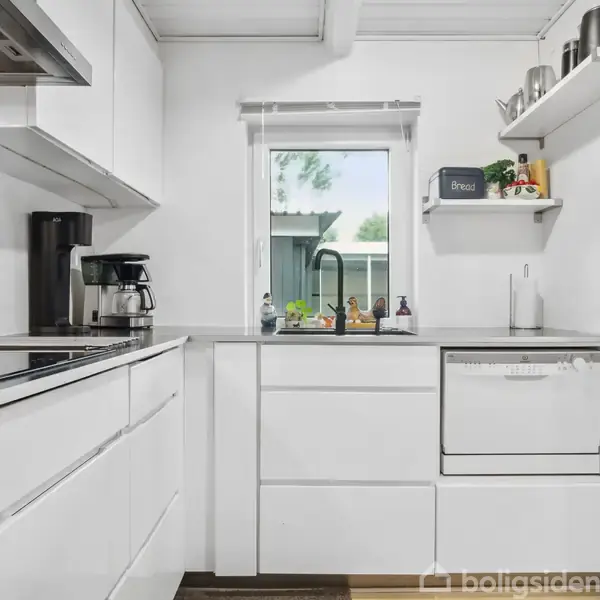 Et hvidt køkken med en kaffemaskine og opvaskemaskine under en vindue. Hylder med krukker og en kasse mærket "Brødkasse." Udenfor ses grønne træer og et hegn.