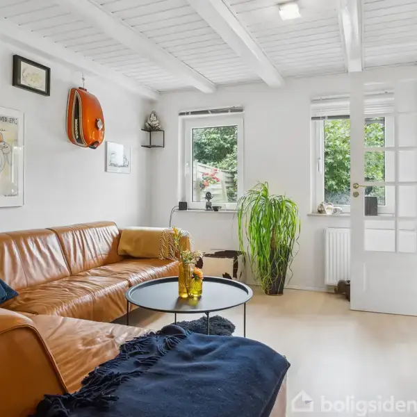 En brun lædersofa står i en stue ved siden af et rundt sofabord med en vase. Lyse trælofter og store vinduer skaber en lys atmosfære med udsigt til grønne planter.