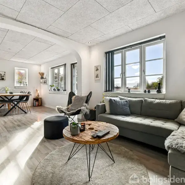 Stue med grå sofa og rundt træbord foran stort vindue, giver naturligt lys. I baggrunden ses spisebord med stole i den åbne spiseafdeling. Vægge har hylder og dekorationer.