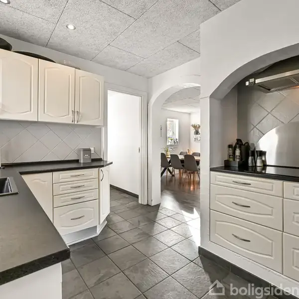 Et moderne køkken med hvide skabe og sorte bordplader strækker sig langs væggene. I baggrunden ses en buegang, der leder ind til en spisestue med et bord og stole.