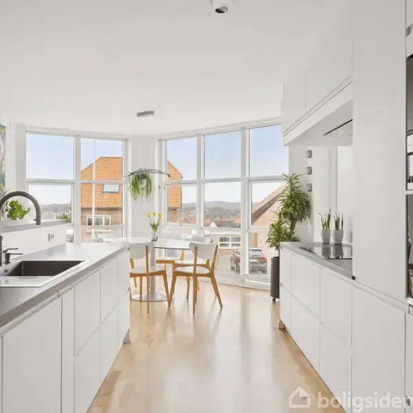 Et moderne køkken med hvide skabe, indbygningsovn og sort bordplade. En stor rund spiseplads er ved gulv-til-loft vinduer med udsigt over tage. Grønne planter pryder rummet.