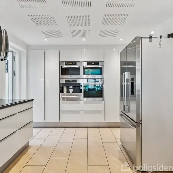 Et moderne køkken indeholder en stor metal køleskab, indbyggede hvidevarer og en sort emhætte, omgivet af hvide skabe. Der er lyse fliser på gulvet og spots i loftet.