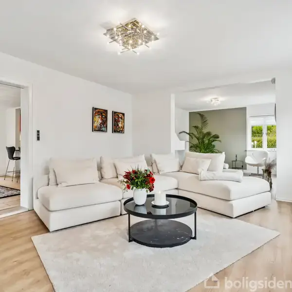 En stor, lys stue med en cremefarvet sofa placeret på et lyst tæppe. Et rundt sofabord står foran sofaen med en blomsterdekoration. Til venstre er der en åbning til spisestuen, der har et spisebord og stole. Rummet har trægulve og hvide vægge med to vægbilleder.