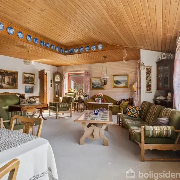 Stue med traditionelle møbler, herunder sofagruppe og spisebord. Vægge prydet af malerier og blå tallerkener. Træloft og store vinduer skaber en lys, varm atmosfære.