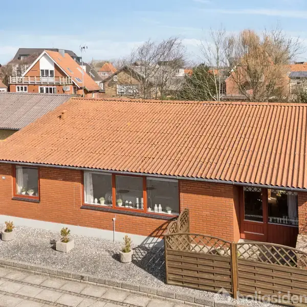 Et rødt murstenshus med rødt tegltag står stille i et beboelsesområde. Foran er en grusbelagt gårdhave med nogle planter. Flere huse kan ses i baggrunden.