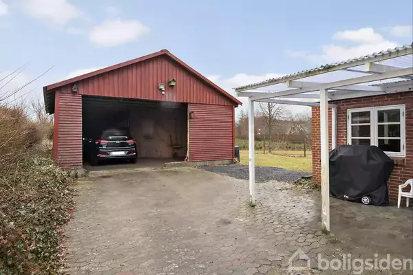 En bil holder i en rød garage ved siden af en beskeden gårdsplads med fliser. Til højre er et murstenshus med en overdækket terrasse og grilludstyr foran en græsplæne.