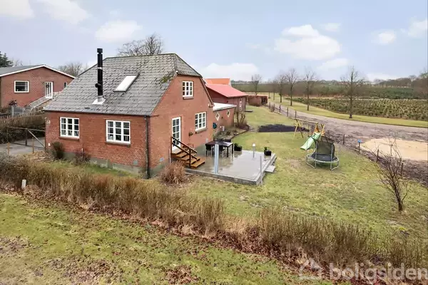 Et rødt murstenshus står stille i en grøn have med terrasse, legeområde og trampolin. Omkring huset er en mark og spredte træer under en blå himmel med få skyer.