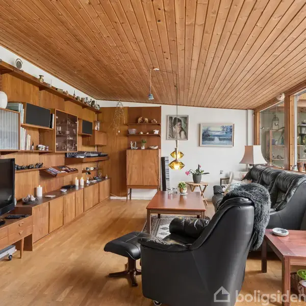 Stue med sort lænestol og sofabord, placeret foran reolsystem med TV og dekorative genstande. Store vinduer til højre giver udsigt. Trægulv og loft skaber varm atmosfære.