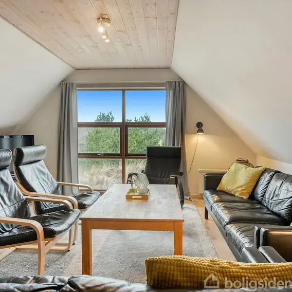 Stue med sorte lædermøbler, to stole og en sofa, omkring et træbord. Vindue med udsigt til natur, trægulv og loft, og dæmpet belysning. Gule puder pynter sofaen.
