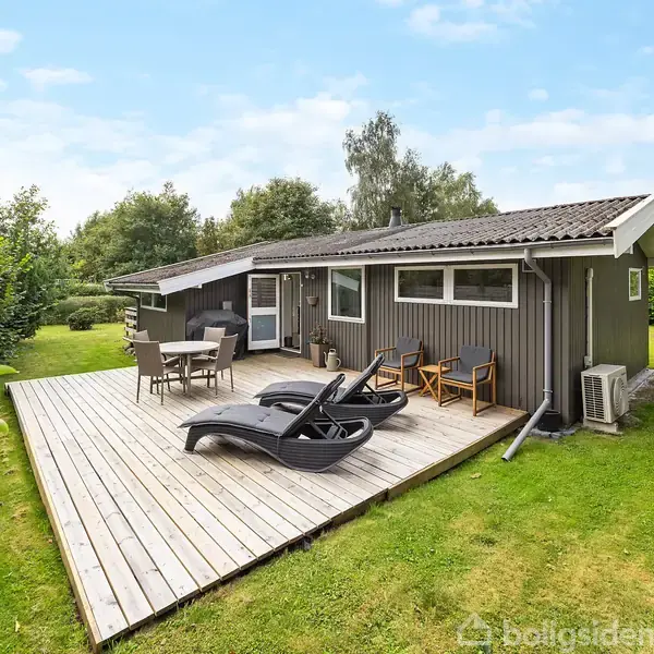 Et sommerhus i træ ligger i en have. Huset har en stor træterrasse med liggestole og havemøbler. Omgivet af græsplæne og grønne træer under en klar blå himmel.