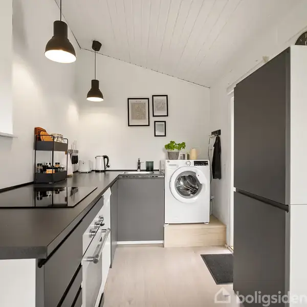 Et moderne køkken med grå og hvide skabe, en vaskemaskine i baggrunden og to hængende lamper. Rummet har minimalistisk indretning med planter og billeder på en hvid væg.