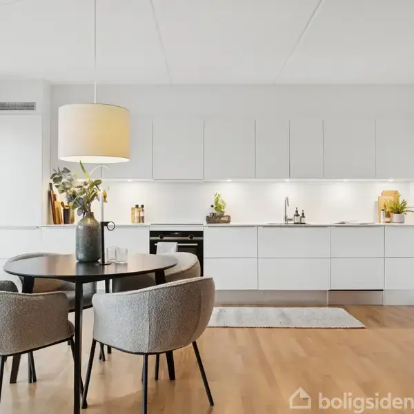 Rundt spisebord med fire polstrede stole, står i moderne, lyst køkken. En stor lampe hænger over bordet. Køkkenet har hvide skabe, træ- og plantepynt, og trælaminatgulv.