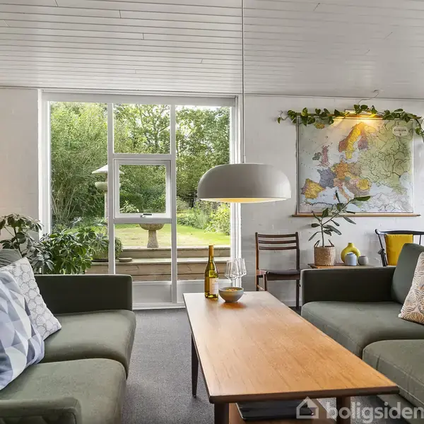 To grønne sofaer omgiver et rektangulært træbord med en vinflaske og glas. Et stort vindue viser en have udenfor. Kort og planter dekorerer væggen.