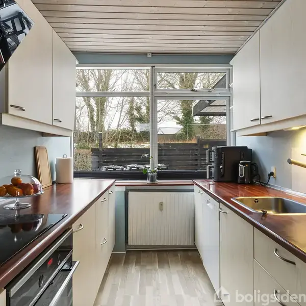 Et lyst køkken med hvide skabe og brun bordplade. Der står køkkenudstyr og appelsiner på bordet. Gennem vinduet ses en have med træer.