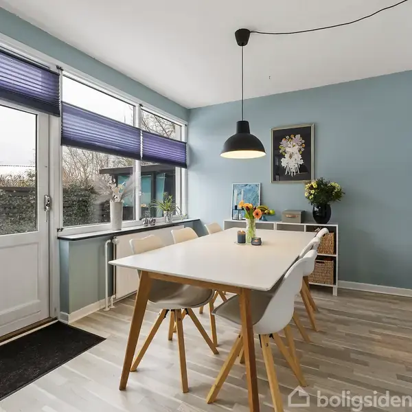 Et spisebord står i et lyst rum med blå vægge. Vinduer med blå persienner fylder venstre side, mens en loftslampe hænger over bordet. Blomster og kunst dekorerer rummet.