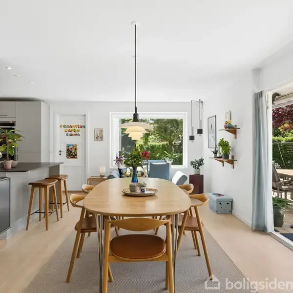 Et moderne spisestueområde med et langt træbord omgivet af stole. En lampe hænger over bordet. Et stort vindue og skydedør bagved giver udsigt til en terrasse og have.