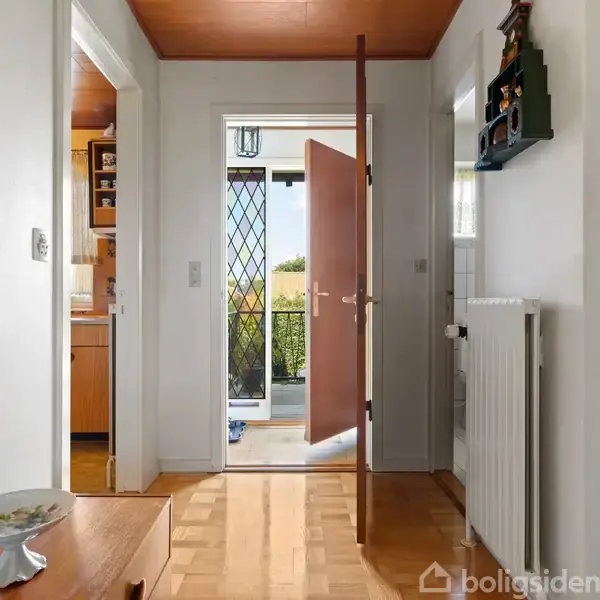 En dør står åben mod en solrig veranda, omgivet af lyse vægge og træmøbler. Køkkenet er synligt til venstre. Gulvet har parketmønster, og en radiator er ved siden af.