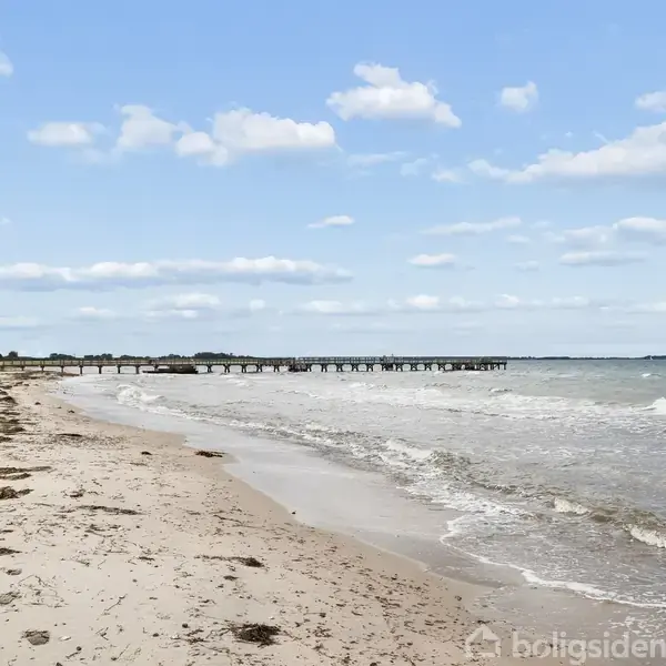 Strand med bølger, tang langs kysten. Bro strækker sig ud i havet. Vindblæst miljø under let skyet, blå himmel.