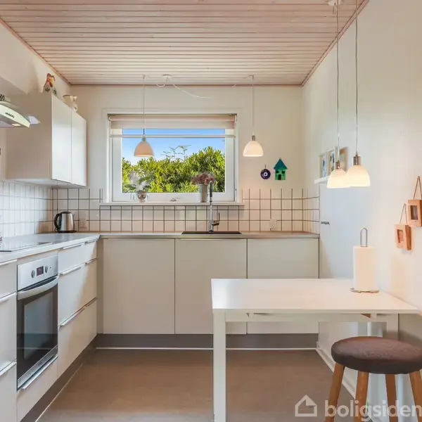 Et moderne køkken har hvide skabe, indbygget ovn, og køleskab. Et hvidt bord med en stol står foran et vindue med udsigt til grønne træer. Pendellamper hænger fra loftet.