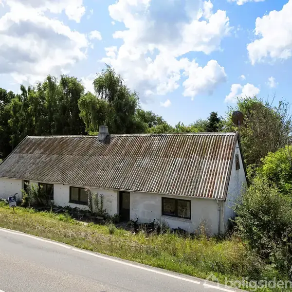 En hvid, ældre hus med skråt tag står ved en landevej. Huset er omgivet af træer og græsarealer under en skyet himmel.