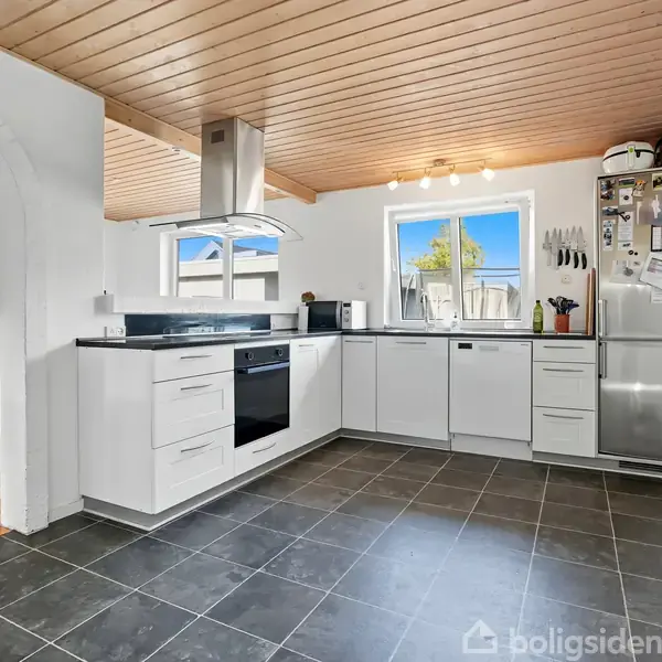 Et moderne køkken med hvide skabe og sorte fliser. Køleskabet er fyldt med magneter og noter. Gennem buen ses en spiseplads med trægulv og mørke stole.