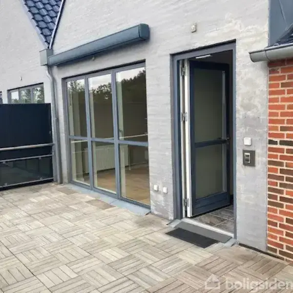 En terrassedør står åben ved siden af store vinduer i en moderne bygning, der har murstensdetaljer og grå vægge. Flisebelagt terrasseområde med glasrækværk omgiver bygningen.