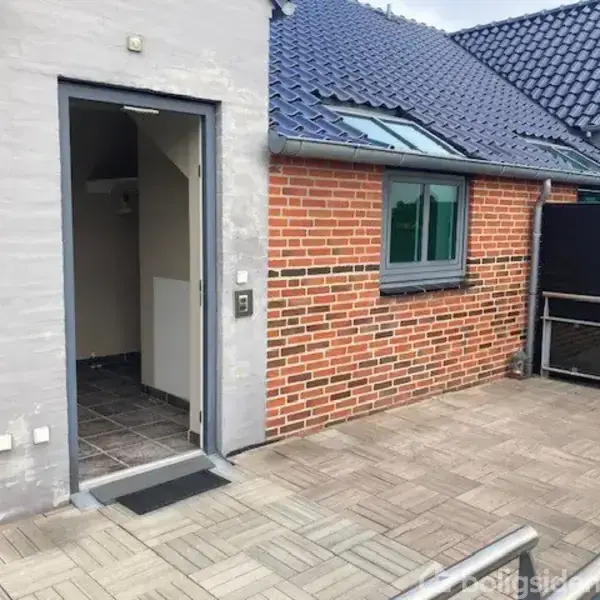 En udendørs terrasse med trægulv. En åben dør fører til et indendørs område med flisegulv. Bygningens facade er af rød mursten med grå detaljer og blå tagsten.