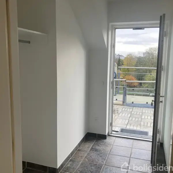 En dør står åben ud til en balkon med glasrækværk. Indendørs er der flisegulv og hvide vægge. Udsigten viser trætoppe og en overskyet himmel.