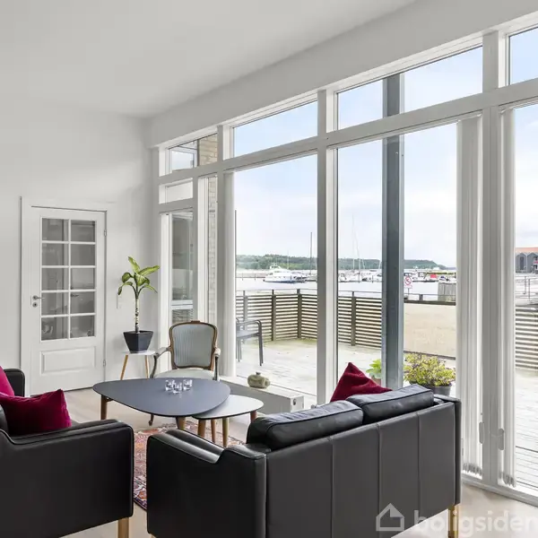 Sorte sofaer står omkring et sofabord i en stue med store vinduer, der åbner op mod en terrasse med udsigt over en marina. En vitrineskab og en plante står ved væggen.