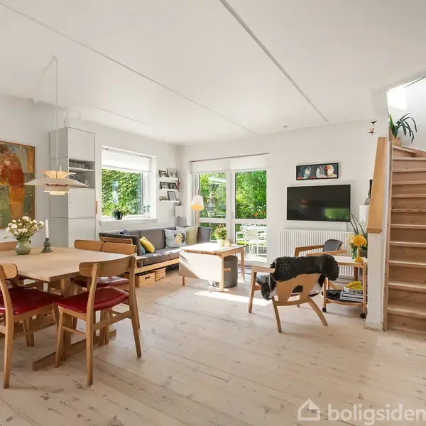 En lys stue med spisebord til venstre, sofa og lænestole foran et stort vindue. Trætrapper fører opad til højre. Bøger og kunst pryder rummet.