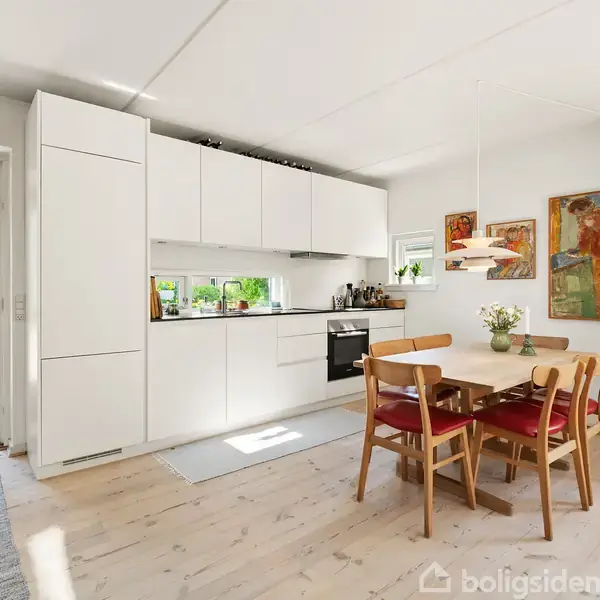 Et lyst køkken med hvide skabe og en indbygget ovn. Et træbord med fire røde stole står ved siden af. Vægge har kunstværker, og en dør fører ud til haven.