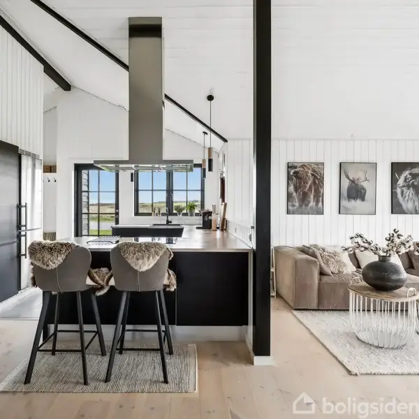 Køkken med sort køkkenø og to barstole med pels. I baggrunden ses en stue med beige hjørnesofa, sofabord og dyremotiver på væggen. Store vinduer giver lysindfald.