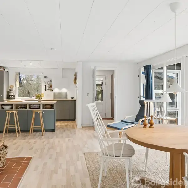 En lys stue-køkken-alrum med brændeovn og kurv med brænde. Rundt spisebord med stole står midt i rummet. Store vinduer og dobbeltdøre fører ud til terrasse.