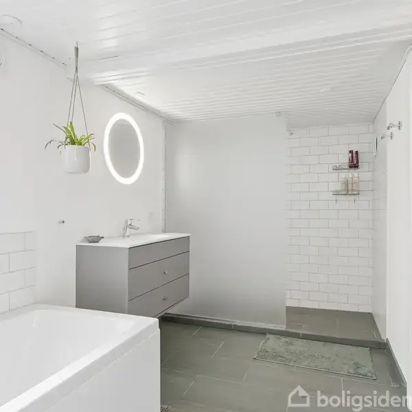 Et lyst badeværelse med badekar, moderne håndvask med skuffer og rund oplyst spejl. Hvide fliser på væggene, grå flisegulv, en hængende plante, og en åben dør til højre.