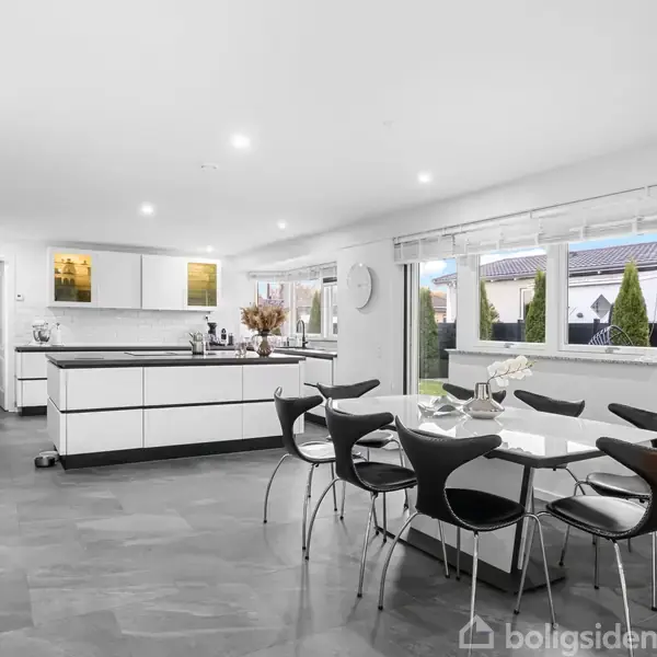 Køkkenet har et moderne design, indeholder en hvid køkkenø og spisebord med sorte stole. Store vinduer giver udsigt til en have. Rummet er lyst og rummeligt med grå gulve.