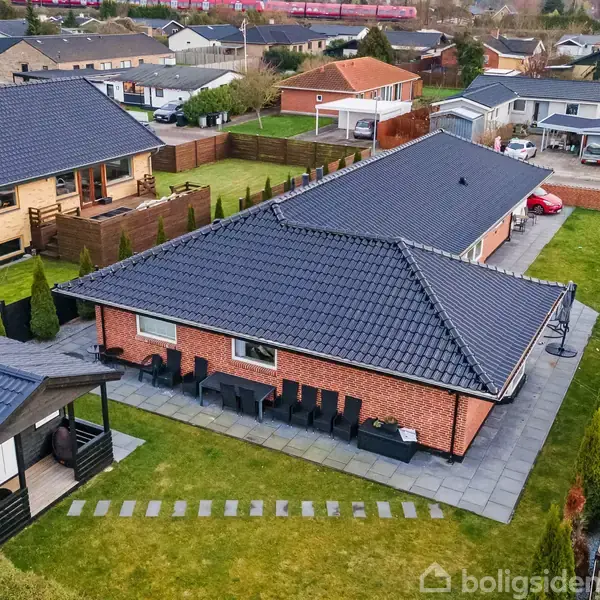 Rødt murstenshus med sort tag omkranser have med flisebelagt terrasse. Lang havebord står udenfor. Omgivet af nabolag med lignende boliger og grønne arealer.