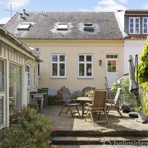 Terrassemøbler står omkring et rundt bord på en gårdsplads omgivet af to bygninger. Området har blomster og planter, mens en grill står til højre op ad en gul murstensbygning.