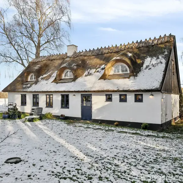 Et hvidt, stråtækt hus med runde vinduer står i et snedækket landskab med nøgne træer og en klar himmel.