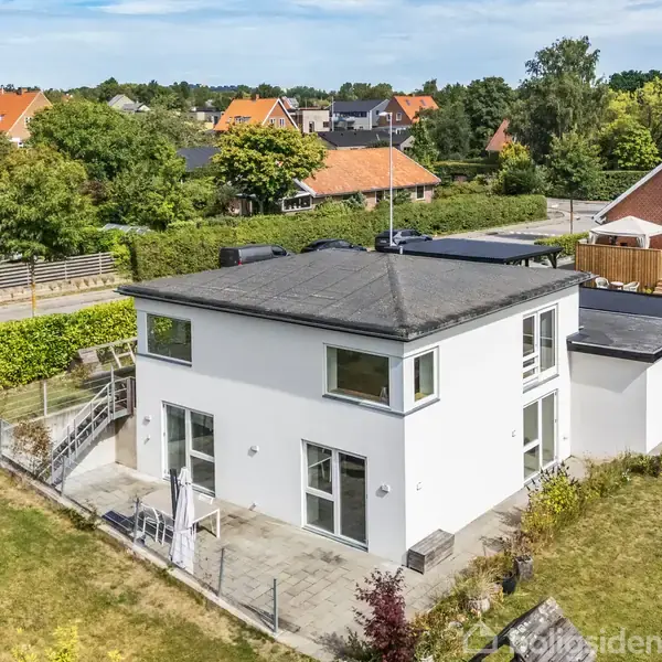 Huset står stille i en frodig have, med en hvid facade og store vinduer. Omkranset af hæk ligger det i et villakvarter med andre rødtage huse.