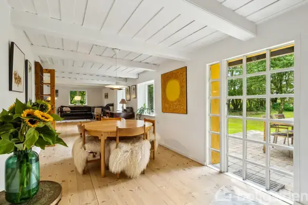 Træbord omgivet af pelsdækkede stole; står i et lyst rum med træloft, vinduer og kunst på væggene; udsigt til terrasse og grøn have gennem glasdør; solblomstdekoration på bordet.