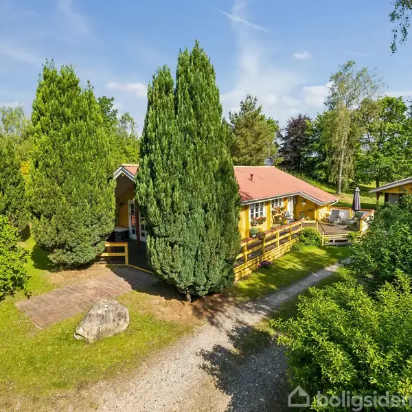 Hus med rødt tag omgivet af høje, grønne træer og frodigt buskads. En stenbelagt sti leder til hoveddøren gennem den grønne have på en solrig dag.