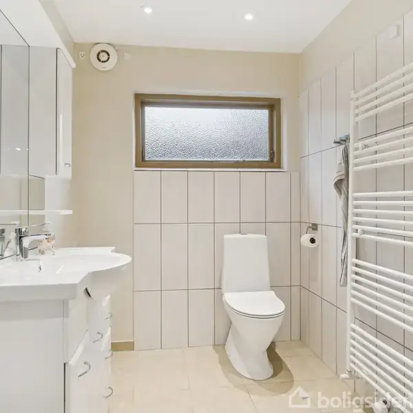 Toilet står mod flisevæg i lyst badeværelse. Til højre hænger et håndklædetørrer. Et spejlskab og håndvask med skuffer er til venstre. Rummet har et lille, frostet vindue.