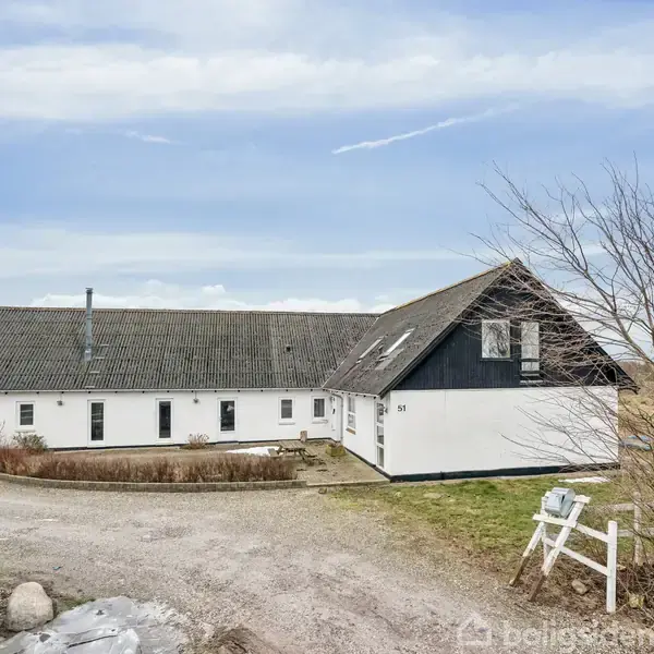 Et stort, hvidt landhus med sort tag står på en grusvej. Der ligger marker og åbne landskaber omkring huset. Nummeret "51" er synligt på bygningens facade.