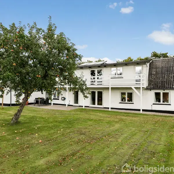 Hvidt hus med sort tag har to etager og balkon, omgivet af grøn græsplæne og træer i en have. Himlen er klar med få skyer.